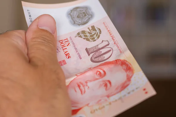 Persons hand giving the Currency of the Singapore - One red ten dollar note spread out on a brown background. Money exchange.