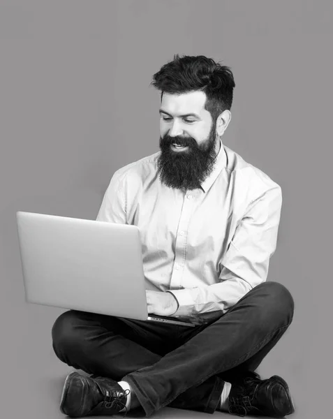 Sonriendo guapo barbudo trabajador portátil. Joven feliz sentado en el suelo con y el uso de la computadora portátil en el fondo rojo. Joven hombre de negocios usando su portátil, pc. Blanco y negro —  Fotos de Stock