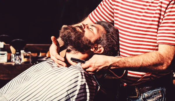 Barber shaving a bearded man in a barber shop. Bearded male in a barber shop while hairdresser shaves his beard with a dangerous razor. Straight razor, barbershop, beard. Vintage straight razor