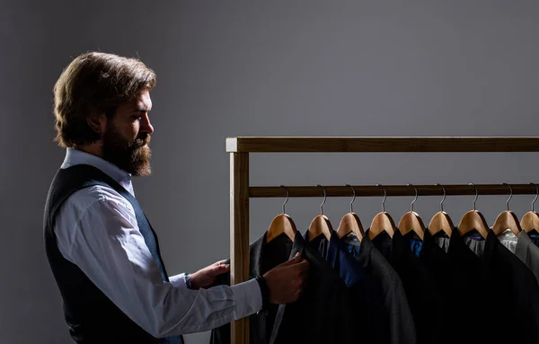 Tailor, tailoring. Stylish mens suit. Male suits hanging in a row. Men clothing, boutiques. Man suit, tailor in his workshop. Handsome bearded fashion man in classical costume suit