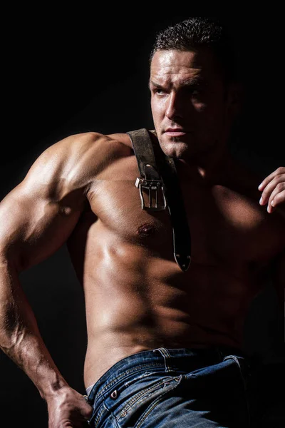 Muscular model sports young man on dark background. Fashion portrait of strong brutal guy. Leather belt, jeans. Sexy torso. Sport workout bodybuilding concept. Sensual mans body