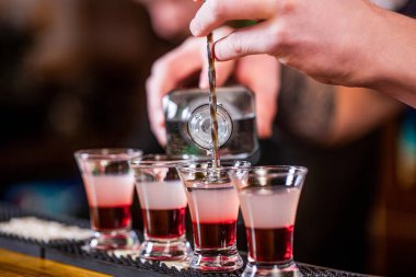 Barmen bardaki küçük bardaklara güçlü alkollü içecekler dolduruyor. Gece kulübünde fondip. Bardakta kırmızı alkolik içkisi. Gece kulübünde kırmızı kokteyl. Barmen kokteyl hazırlıyor.