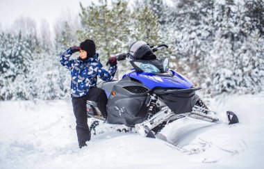 Çocuk, karlı bir yolda, ormanda mavi bir kar aracına biniyor. Kış manzarasında kar arabası süren bir çocuk. Karlı bir havada, ormanda, dağların tepesinde küçük bir çocuk gülümsüyor..