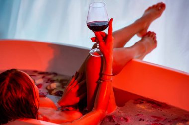 Gül yapraklarıyla banyo yaparken elinde bir bardak şarap tutan bir kadın. Köpüklü bir küvette şarap ve şampanya içen güzel bir kadın. Güzel seksi kız, spa tedavisinde rahatla..