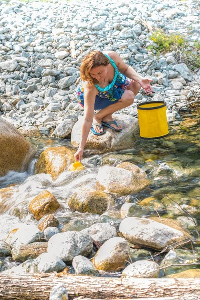 Woman pours with cup of water in a bucket from wild stream