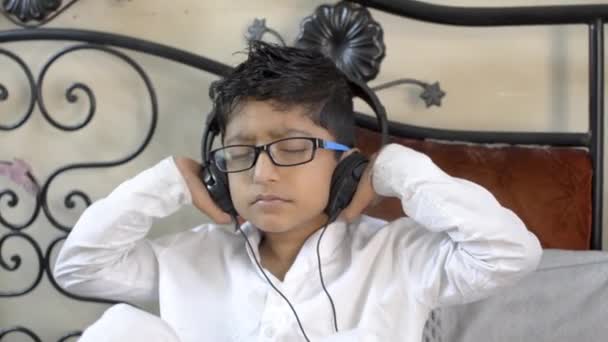 小さなインドのアジアの白人の少年は ヘッドフォンの閉じる目とリビングルームの側面のビューに座って耳に手を楽しんで音楽を聴いて眼鏡を着用 — ストック動画