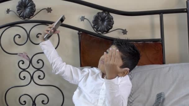 可爱的可爱的小印度亚洲白人男孩孩子采取有趣的自拍在手机或智能手机相机与有趣的表情在卧室特写拍摄侧视图 — 图库视频影像