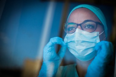 Pencerenin yanında, maske ve gözlük takan genç bir kadının renkli portresi. Coronavirus Covid-19 pandemisi için bir resim..