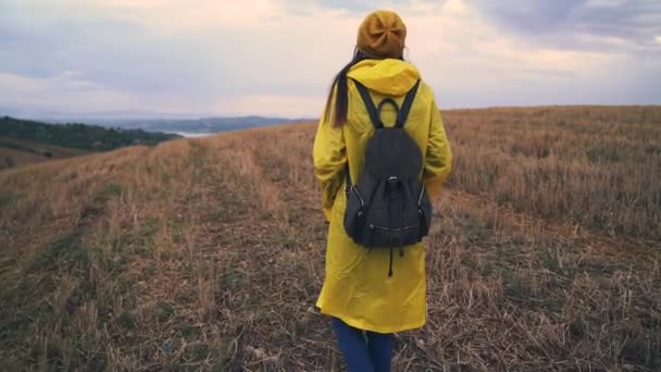 Мандрівник жінка ходить по полю. Дівчина в жовтому плащі з рюкзаком — стокове відео