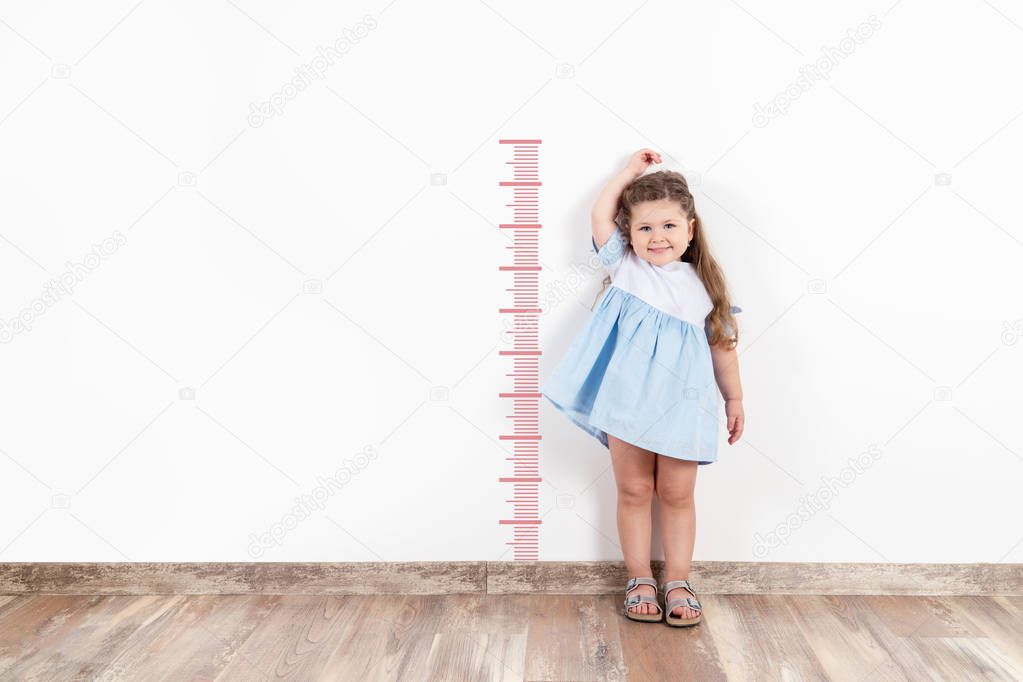  Little blond girl measuring height on white wall.