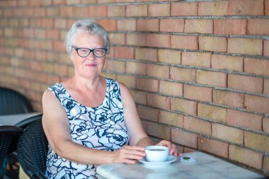 Yaşlı kadın kameraya bakıyor ve kahveyle gülümsüyor.