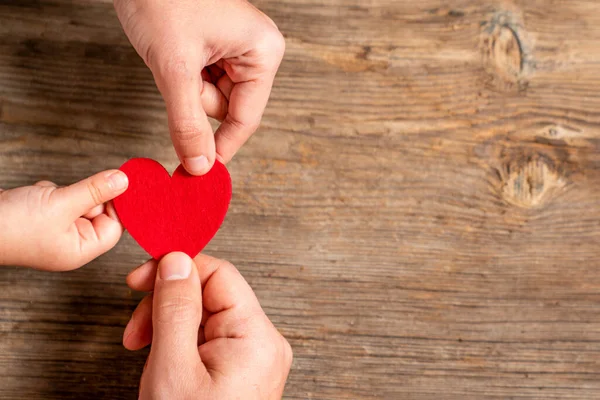 Οικογενειακά χέρια κρατώντας κόκκινη καρδιά σε ξύλινο φόντο. Αγάπη, δωρεά, φιλανθρωπία, έννοια της υγείας. — Φωτογραφία Αρχείου