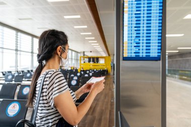 Virüs korumalı genç bir kadın havaalanındaki bilgi panosuna bakıyor. Corona virüsü salgını.