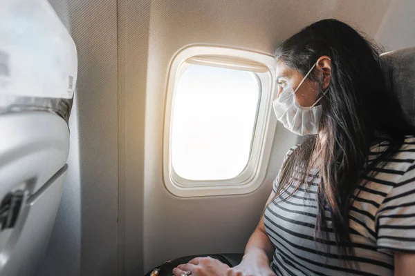 保護マスクの若い女性が飛行機に座って窓からの眺めを見ています。コロナウイルス発生. — ストック写真