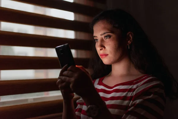 Молодая и серьезная женщина ищет смартфон в темной комнате . — стоковое фото