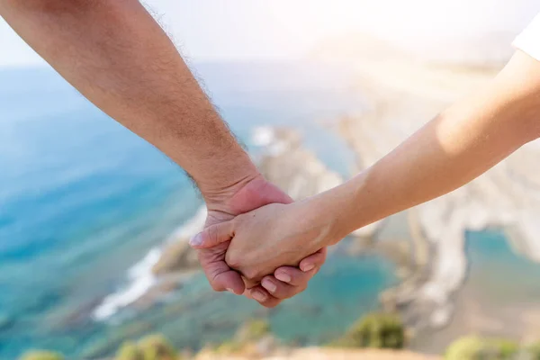 Zwei Hände halten sich mit dem verschwommenen Sonnenlicht und der schönen Aussicht auf eine Klippe und das Meer. — Stockfoto