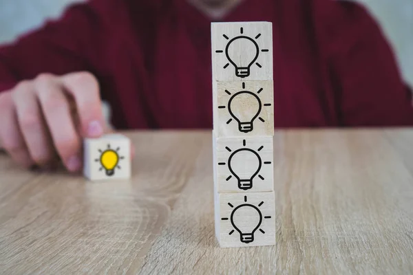 手在木制桌子上挑了一个黄色灯泡符号的木制立方体。新概念、创新和解决方案概念. — 图库照片