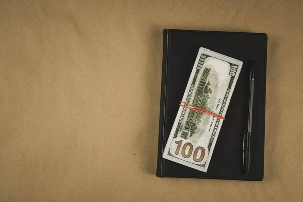 Un carnet noir, un stylo et des billets en espèces en dollars sur fond de bois concept de gestion ou de planification financière, gagner de l'argent en écrivant à la pige — Photo