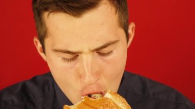 Kırmızı arka planda özenle hamburger yiyen bir adam.