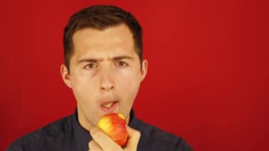 Kırmızı arka planda duran adam özenle bir Apple yiyor.