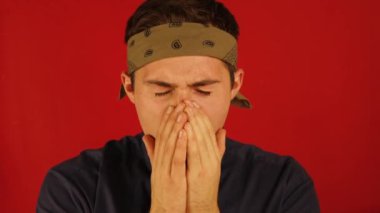 Bandanalı bir adam esniyor ve gözlerini kırmızı arka plana sürtüyor.