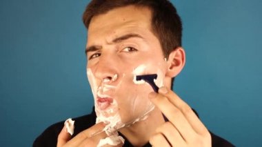 Genç bir adam mavi arka planda dikilirken tıraş olur.