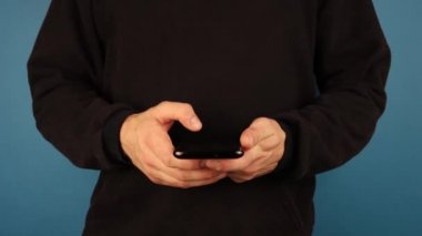 Siyah kazaklı bir adam mavi arka planda kameraya bakarken telefon kullanıyor.