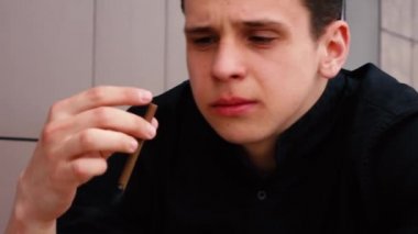 Siyah gömlekli bir adam sokakta sigara içiyor.