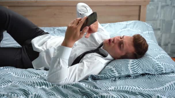 Ένας κουστουμάτος είναι ξαπλωμένος στο κρεβάτι χρησιμοποιώντας το τηλέφωνό του. — Αρχείο Βίντεο