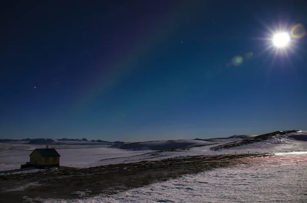 The moon with the rays and the Aurora in Iceland s blue winter sky over an Icelandic house that stands on a lava field covered with snow.