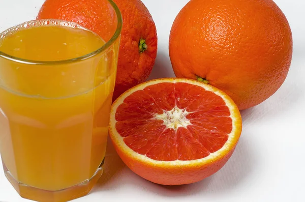 Un vaso de jugo de naranja sobre el fondo blanco se encuentra junto a dos naranjas enteras y naranja roja en rodajas —  Fotos de Stock