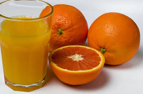 Un vaso de jugo de naranja sobre el fondo blanco se encuentra junto a dos naranjas enteras y naranja roja en rodajas —  Fotos de Stock