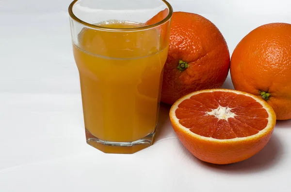 Un vaso de jugo de naranja sobre el fondo blanco se encuentra junto a dos naranjas enteras y naranja roja en rodajas —  Fotos de Stock