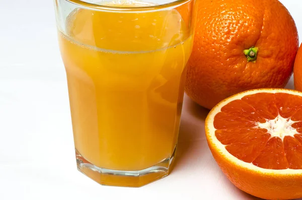 Un vaso de jugo de naranja sobre el fondo blanco se encuentra junto a dos naranjas enteras y naranja roja en rodajas —  Fotos de Stock