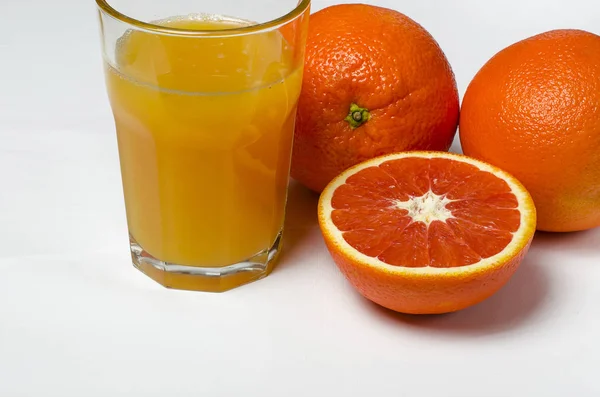 Un vaso de jugo de naranja sobre el fondo blanco se encuentra junto a dos naranjas enteras y naranja roja en rodajas —  Fotos de Stock