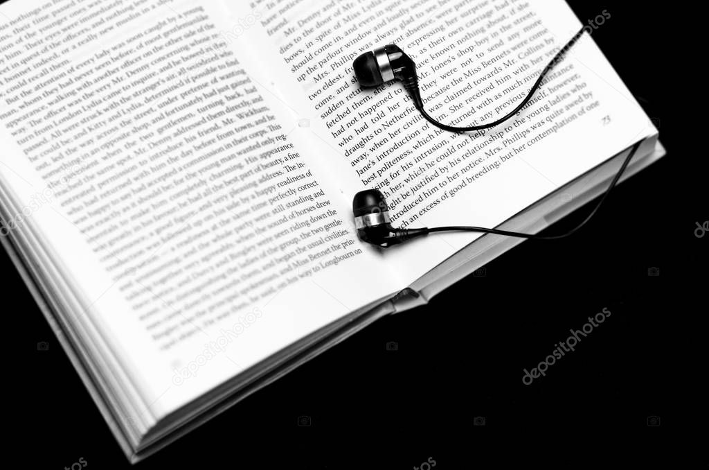 Open ready for reading the book lies on a black background close to black headphones. Book and ear plugs