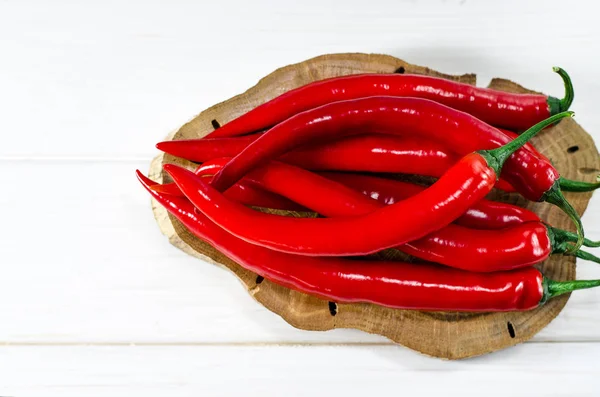 Beaucoup de piments rouges reposent sur un panneau brun ovale, qui se tient sur une table en bois blanc — Photo