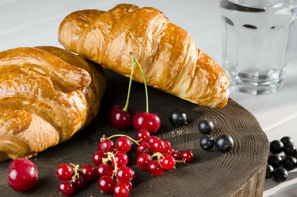 Fresh ruddy croissants with berries lie on a wooden table next to fresh black currant berries, red currants, cherries — Stock Photo, Image