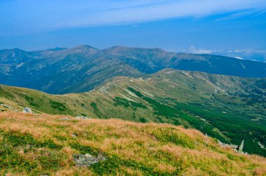 Yüksek dağ sırası bulutlarla mavi gökyüzüne karşı yeşil ve sarı otlarla büyümüş.