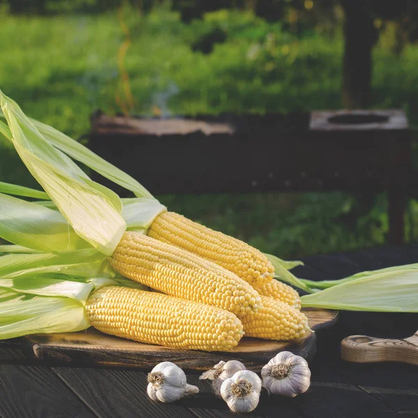 Yellow juicy corn with green leaves lies on a wooden table in the summer garden against the backdrop of a grill — Stock Photo, Image
