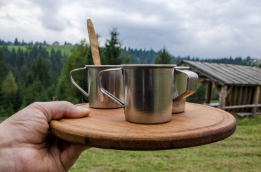 Adamın eli, orman ve dağların arka planında tahta bir tahta üzerinde üç fincan kahve içeren bir tepsi tutar.