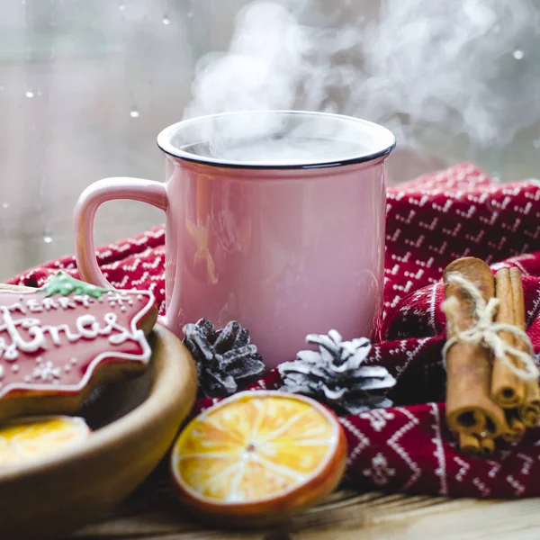 Чашка горячего чая стоит на деревянном столе рядом с деревянной тарелкой, на которой пряничное печенье из оранжевых ломтиков на фоне окна с капельками воды — стоковое фото