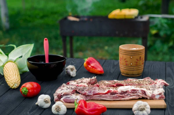 Pork ribs in a marinade lie on a wooden table in the garden next to pepper, garlic, corn on the grill background — Stock Photo, Image