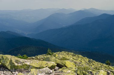 Yeşil likenlerle kaplı büyük taşlar, eski, yüksek sıradağların arka planında dağın tepesinde yatıyor.