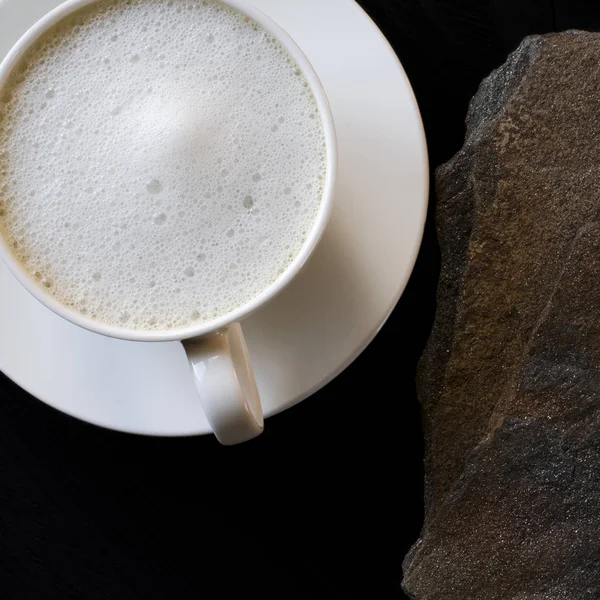 Una tazza di caffè bianco con una spessa schiuma si trova su un tavolo nero accanto a una pietra selvatica multicolore — Foto Stock