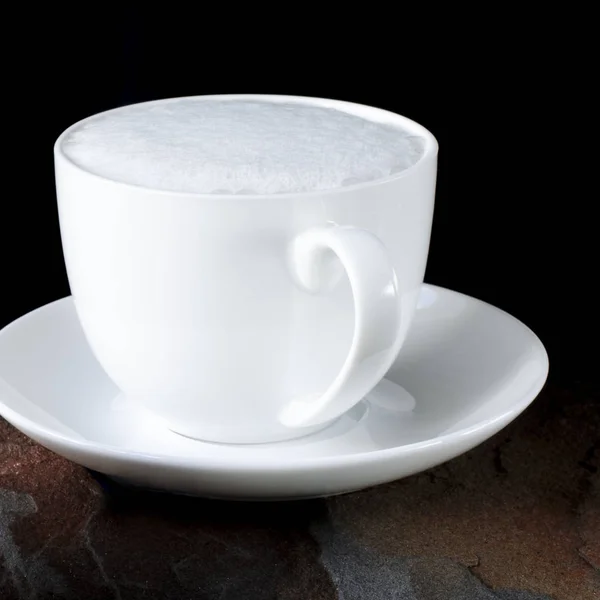 Una tazza di caffè bianco con una spessa schiuma si trova su un tavolo nero accanto a una pietra selvatica multicolore — Foto Stock