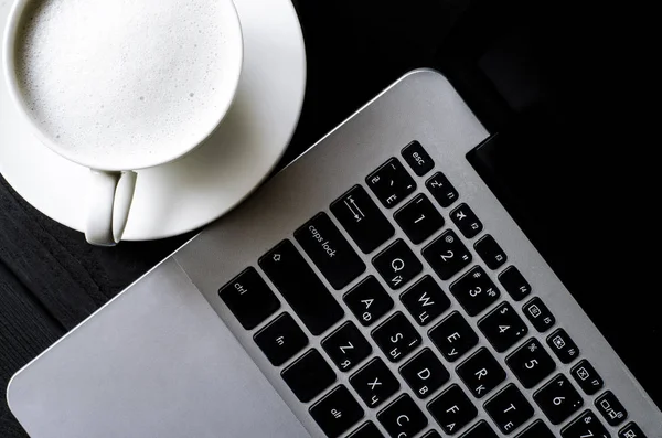 The laptop stands on a black wooden table next to a cup of coffee with foam