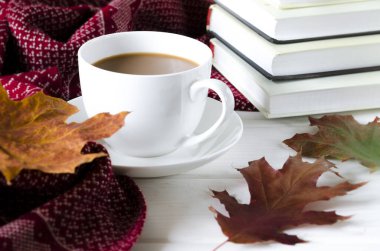 Yuvarlak beyaz bir fincan ve çay tabağı kitapların ve sarı sonbahar yapraklarının yanında beyaz bir ahşap masanın üzerinde duruyor.