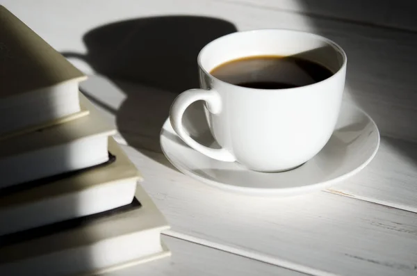 A round white cup of coffee stands on a white saucer on a white wooden table next to the shadow of the cup