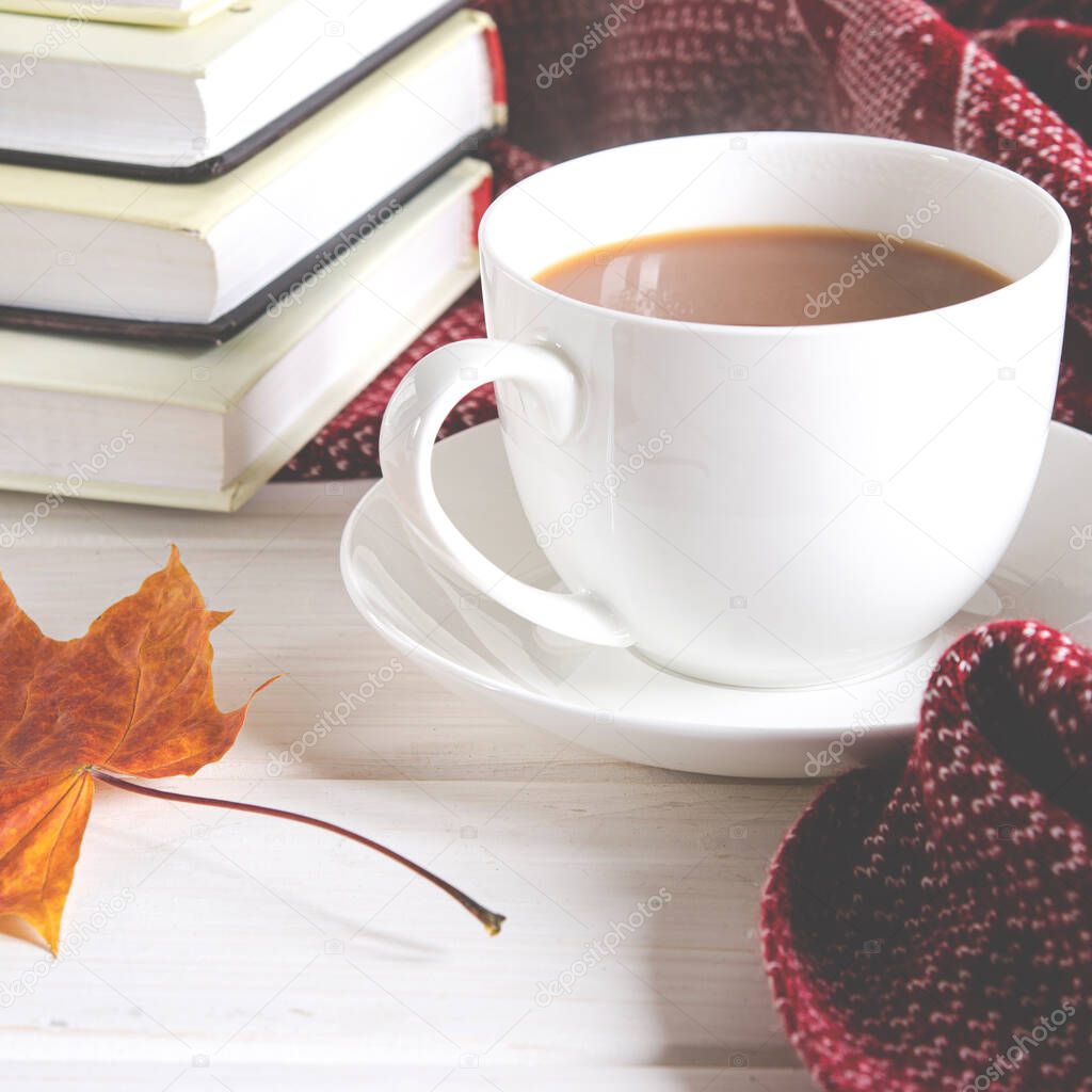 A cup of coffee with milk stands on the table next to books and a warm wool scarf. Close-up